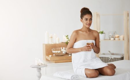 Young beautiful african girl in towel smiling holding glass with tea sitting in spa salon.