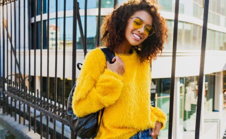 Outdoor bright portrait of happy African girl with back pack  and standing on urban background. Wearing yellow sweater and sunglasses. Pretty student enjoying sunny spring day.